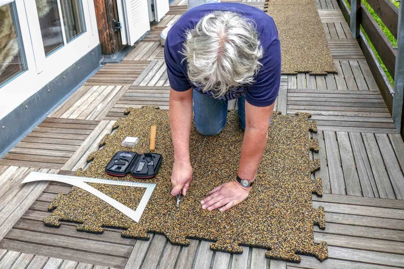 Auf einem Balkon liegen verwitterte Fliesen aus Holz. Die Balkonfläche wird durch das Auflegen von Warco Balkonplatten im Farbdesign Rattan Lounge saniert. Ein Mann schneidet eine Platte passend zum Verlegen zu.