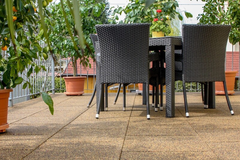 Auf einer Terrasse, die mit quadratischen Terrassenplatten in einem erdfarbenen Farbdesign ausgelegt ist, steht eine Sitzgruppe. Entlang des verzinkten Geländers sind Citrusbäume aufgestellt.