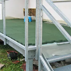 Aus Metall gefertigte Terrasse mit grünen Platten, die an ein Haus angrenzt. Eine kleine Treppe führt zwei Stufen hinauf. Der Unterbau besteht aus Gitterrost.