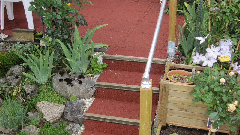 Kleine rote Treppen im Garten die sich durch Pflanzen hoch auf ein Terrasse zieht.