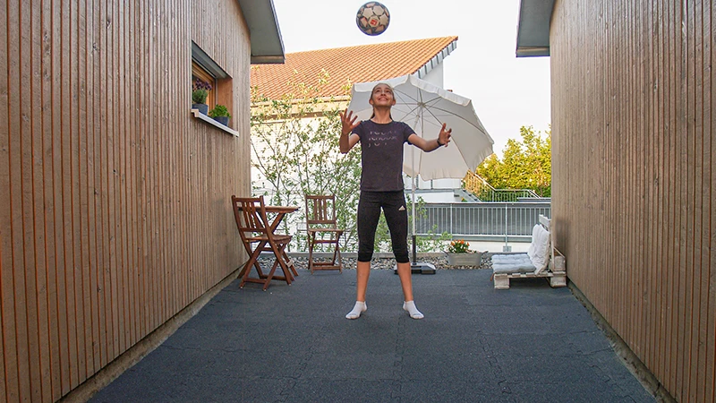 Ein Mädchen spielt Ball auf einer Terrasse zwischen zwei holzverkleideten Gebäudewänden. Der Bodenbelag besteht aus schwarzen Terrassenplatten aus Gummigranulat. Die Terrasse bietet eine robuste und trittelastische Fläche, die für sportliche Aktivitäten geeignet ist. Im Hintergrund steht ein Sonnenschirm und eine kleine Sitzgruppe aus Holz, die eine einladende Atmosphäre schaffen.