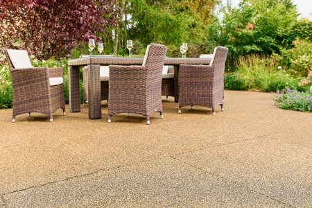 Blick auf eine sonnige Terrasse mit beigebraunen Terrassenplatten von WARCO. Der Bodenbelag aus pu-gebundenem Gummigranulat ist rutschfest, wasserdurchlässig und sorgt für hohen Gehkomfort. Ein stabiler Gartentisch mit geflochtenen Stühlen und gefüllten Weingläsern lädt zum Entspannen ein. Rundherum blühen üppige Sträucher und bunte Blumen, die eine naturnahe Atmosphäre schaffen. Die elastische Oberfläche ist frostbeständig, dämpft Trittgeräusche und bleibt formstabil.