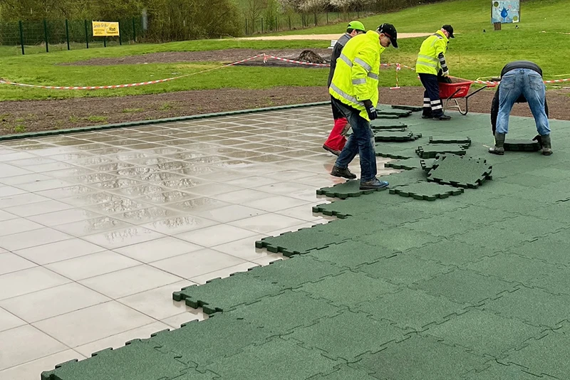 Eine Gruppe von Arbeitern beim Verlegen von grünen WARCO Outdoor Klickfliesen bei Regen auf einer gekachelten Fläche im Freien.