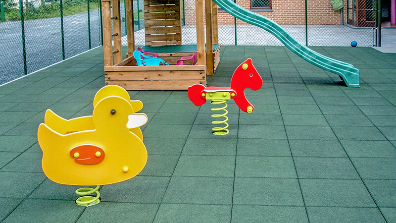 lick auf einen Kinderspielplatz, dessen Spielfläche komplett mit grünen Fallschutzplatten von WARCO ausgelegt wurde. Auf dem Kinderspielplatz stehen verschiedene Spielgeräte.
