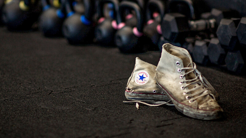 Auf schwarzen Fitnessmatten mit Puzzlesystem von WARCO steht ein Paar stark abgenutzter Converse ALL STARS Sneakers. Den Hintergrund bildet eine lange Reihe von Hanteln. 