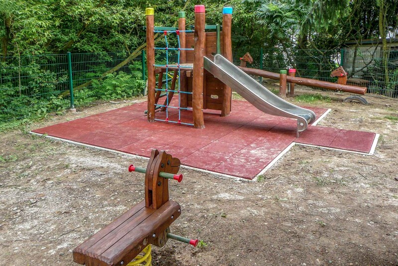Auf einem neu angelegten Kinderspielplatz befinden sich drei Spielgeräte. Im Hintergrund eine Wippe, in der Mitte ein kleiner Kletterturm mit Rutsche und im Vordergrund eine Federwippe. Um den Kletterturm herum sind Fallschutzplatten verlegt.