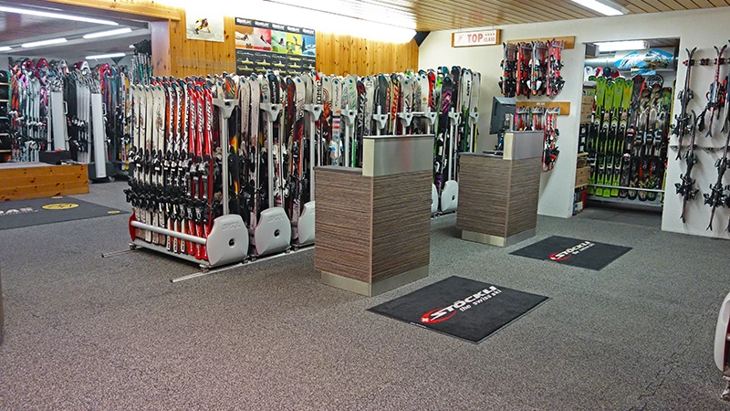 Hinter zwei Theken im Schigeschäft Gaby Sports stehen Ständer mit verschiedenen Schimodellen. Der Boden ist mit Indoor-Klickfliesen von WARCO im Farbton Rattan Lounge belegt.