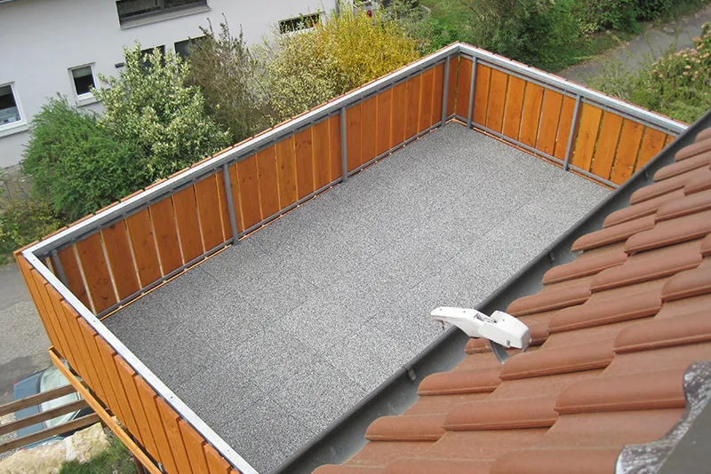 Graue Balkonplatten liegen auf mit Hozlgeländer gesichertem Balkon.  Der Blick fällt vom Dach in den Hof, in dem Büsche und Bäume wachsen.