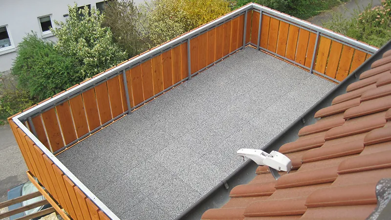 Graue Balkonplatten liegen auf mit Hozlgeländer gesichertem Balkon.  Der Blick fällt vom Dach in den Hof, in dem Büsche und Bäume wachsen.