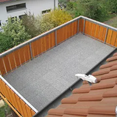 Graue Balkonplatten liegen auf mit Hozlgeländer gesichertem Balkon.  Der Blick fällt vom Dach in den Hof, in dem Büsche und Bäume wachsen.