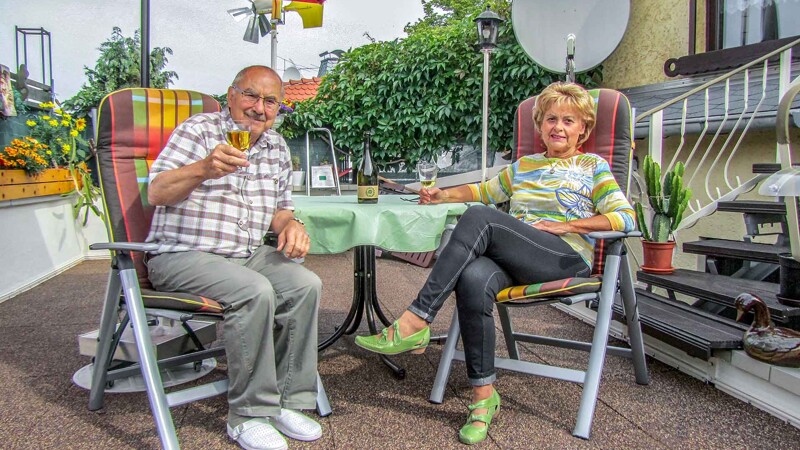 Zufrieden sitzt ein Ehepaar auf der erfolgreich mit WARCO Terrassenplatten im Farbdesign Bast und Borke sanierten Dachterrasse und prostet mit einem Glas Wein vom Weingut Sommer in Hambach in die Kamera.