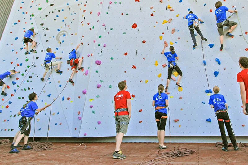 Das Bild zeigt eine Gruppe von Kindern und Jugendlichen, die in einer Indoor-Kletterhalle an einer Kletterwand mit Seilsicherung klettern. Die Teilnehmer tragen blaue und rote T-Shirts und sind mit Klettergurten gesichert. Sie klettern an einer großflächigen Wand, die mit bunten Klettergriffen in verschiedenen Formen und Farben versehen ist. Der Boden in der Kletterhalle ist mit roten Fallschutzplatten von WARCO ausgelegt, die an den Kanten ineinandergreifen und eine stabile, stoßdämpfende Oberfläche bilden. Diese Fallschutzplatten bieten optimalen Schutz und sind ideal für Bereiche mit intensiver körperlicher Aktivität, wie Kletter- und Boulderhallen. Die roten Platten sind nicht nur ein sicherheitsrelevantes Element, sondern auch ein visuell ansprechendes Merkmal der Kletterhalle.