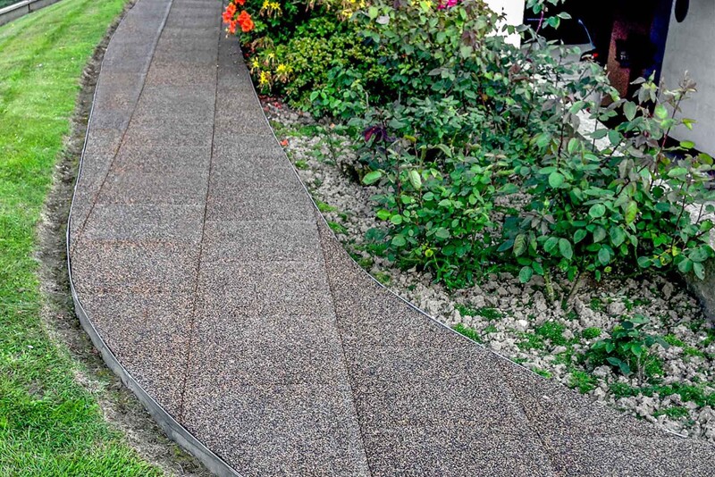 Ein geschwungen gestalteter Gartenweg aus Gummigranulat-Gehwegplatten von WARCO im Farbdesign Bast und Borke führt am Haus vorbei durch den Garten.