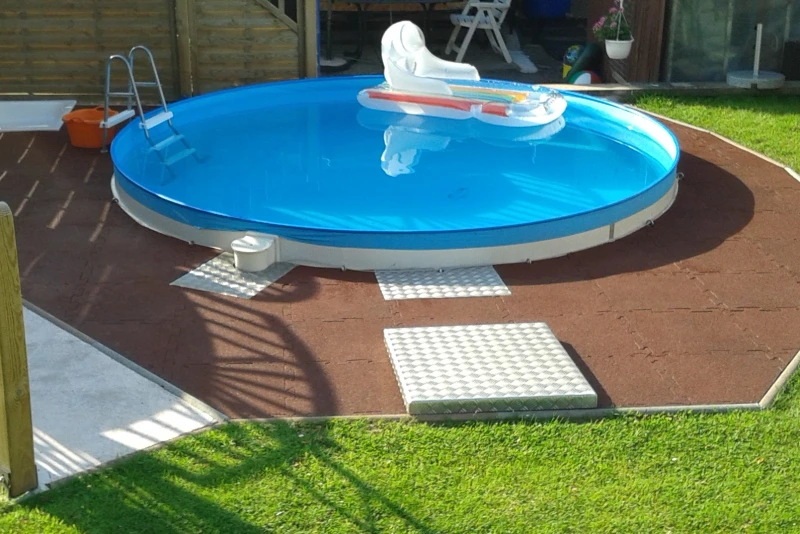 Ein eigener Pool im Garten lässt sich auch auf einem kleinen Grundstück und auch mit geringem finanziellen Aufwand realisieren. Damit das Umfeld um den Pool nicht matschig wird, der Boden aber angenehm zu begehen ist und Fallschutz bietet, wurden Poolfliesen von WARCO verlegt. runder Pool mit braunem Warcobelag.