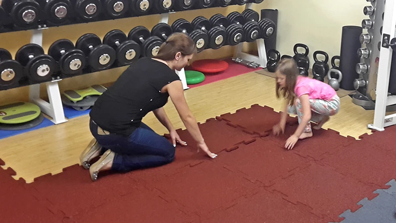 Das Bild zeigt eine Frau und ein junges Mädchen beim Verlegen von WARCO-Fitnessbodenplatten in einer Homegym. Die Bodenplatten sind rot und haben eine ineinandergreifende Schwalbenschwanzverzahnung, die eine einfache und sichere Montage ermöglicht. Im Hintergrund sind mehrere Hanteln, Kettlebells und andere Fitnessgeräte zu sehen, die den Raum als gut ausgestattetes Fitnesszimmer kennzeichnen. Die Frau und das Mädchen arbeiten zusammen, um die Platten genau auszurichten und so eine stabile und rutschfeste Oberfläche zu schaffen. Diese Art von Fitnessboden ist für seine Langlebigkeit bekannt und wird häufig in Fitnessstudios verwendet, um eine sichere und komfortable Trainingsumgebung zu gewährleisten.