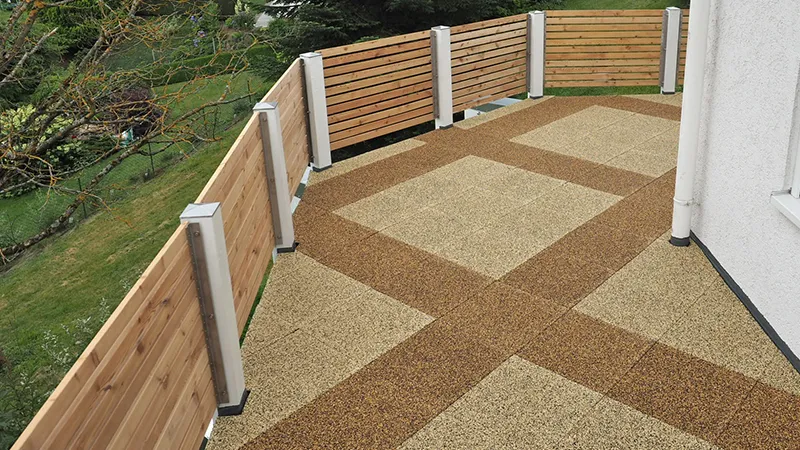 Balkon mit hellbraun dunkelbraun gemusterten Platten. Das hölzerne Geländer passt farblich zu den Balkonplatten. Der Blick fällt in den Garten mit gemähtem Rasen.