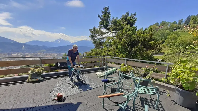 Großer Balkon mit grauen Balkonplatten und Blick in die Berge. Ein Mann hat einen kleinen Tisch aufgebaut und arbeitet an der Verlegung. Zwei grüne Stühle dienen als Ablage.