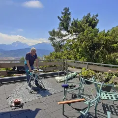 Großer Balkon mit grauen Balkonplatten und Blick in die Berge. Ein Mann hat einen kleinen Tisch aufgebaut und arbeitet an der Verlegung. Zwei grüne Stühle dienen als Ablage.