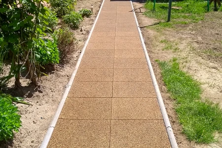 Ein gerader Gartenweg aus quadratischen Gummiplatten mit seitlicher Bordsteineinfassung führt durch das Beet. Die braunen Platten aus Gummigranulat bilden eine pflegeleichte, wetterbeständige und rutschhemmende Oberfläche. Der Weg sorgt für sauberes, sicheres Begehen bei jedem Wetter und grenzt die bepflanzten Flächen klar von der Rasenfläche ab. Ideal für Gärten, Höfe und Zugänge.