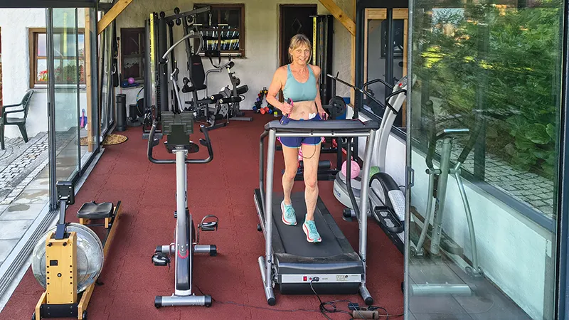 Modernes Homegym mit Glasfront: Eine Frau trainiert auf dem Laufband, der elastische WARCO Fitnessboden schützt Gelenke, dämpft Geräusche und bietet rutschfeste Trittsicherheit – ideal für intensives Training mit Geräten oder Hanteln im privaten Studio.