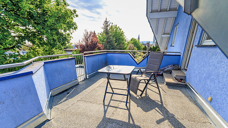 Blau bemaltes Haus mit braunen Balkonplatten. Ein Stuhl und ein Tisch stehen in der Sonne. Große grüne Bäume ragen über den Balkon hinaus.
