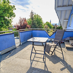 Blau bemaltes Haus mit braunen Balkonplatten. Ein Stuhl und ein Tisch stehen in der Sonne. Große grüne Bäume ragen über den Balkon hinaus.