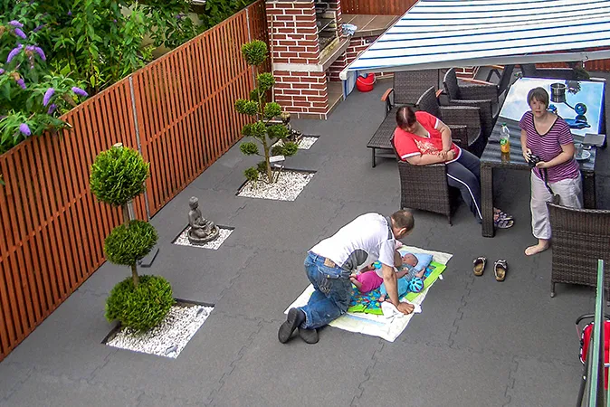 Eine junge  Familie hat sich ihre Dachterrasse als Wohlfühloase mit Sitzgruppe, Grill, Bonsai und dämmendem Terrassenbelag von WARCO im Farbdesign betongrau gestaltet. Das Baby der Familie liegt, nur durch eine Kinderdecke getrennt, direkt auf dem elastischen Terrassenbelag.